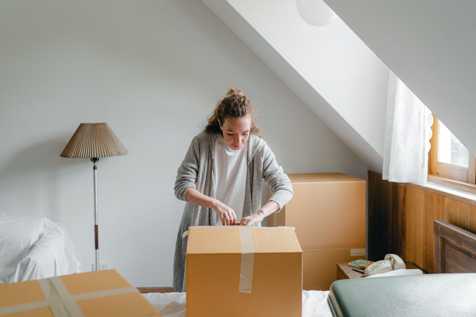 A woman opens a moving box in a bright attic bedroom.