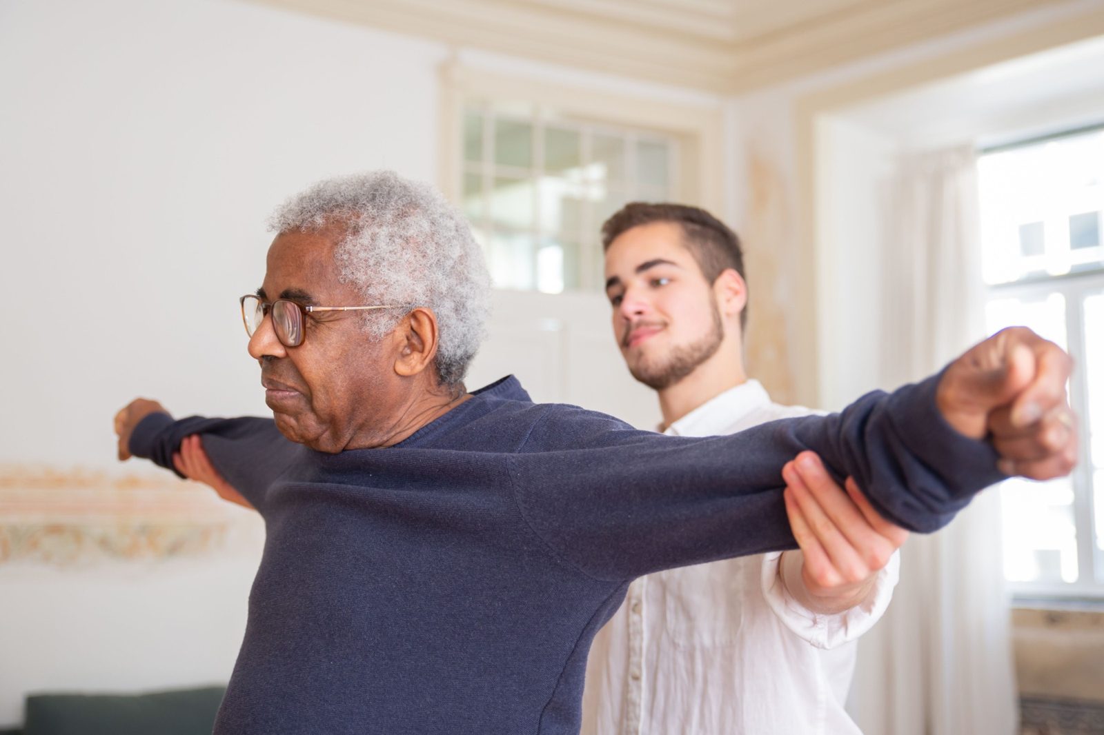 An older man stretches his arms out while a therapist guides the movement in his living room.