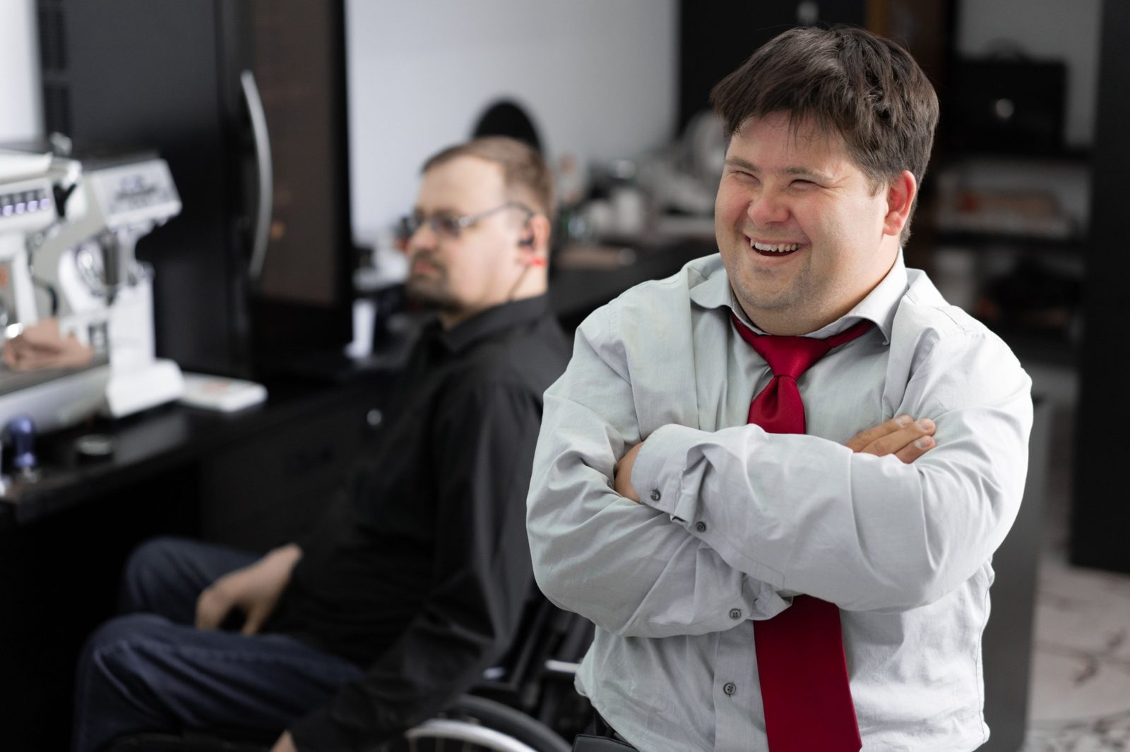 A man in a shirt and tie laughs in his workplace, a colleague at a desk behind him.
