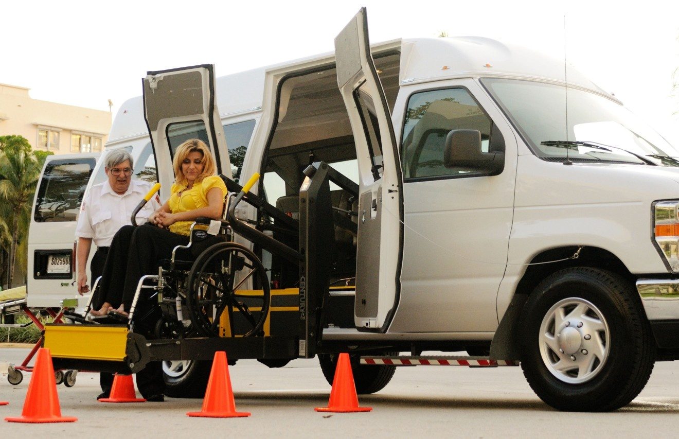 A woman in a wheelchair rides a van's lift up to the door while the driver stands beside her.